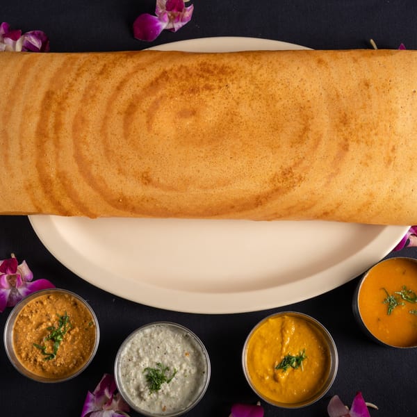 A long, golden-brown dosa on a white plate, surrounded by four small bowls of colorful chutneys.