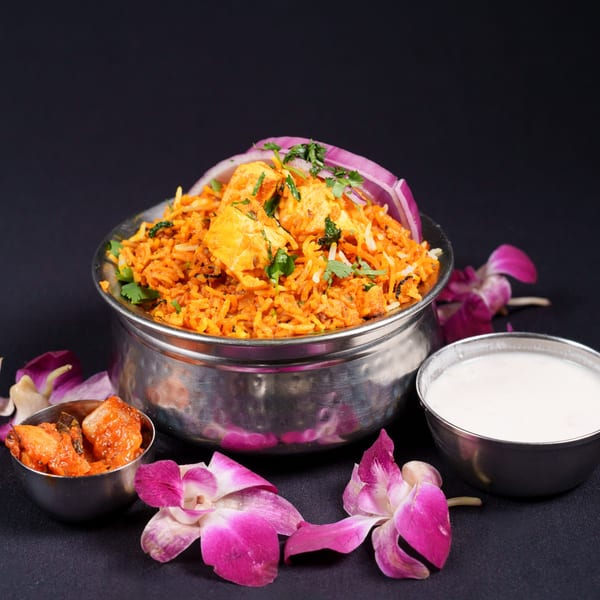 Paneer biryani in a metal bowl, with raita, pickle, and purple orchids on a dark background.