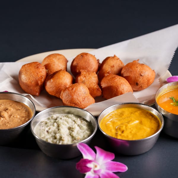 Golden-brown fried fritters on a white paper-lined plate, surrounded by four bowls of colorful dipping sauces.