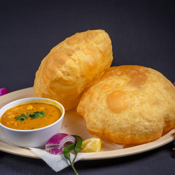 Two golden-brown puri breads with a side of chana masala, red onions, green chilies, and lemon.