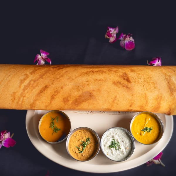 Golden dosa roll with four small bowls of colorful chutneys and sambar, garnished with orchids.